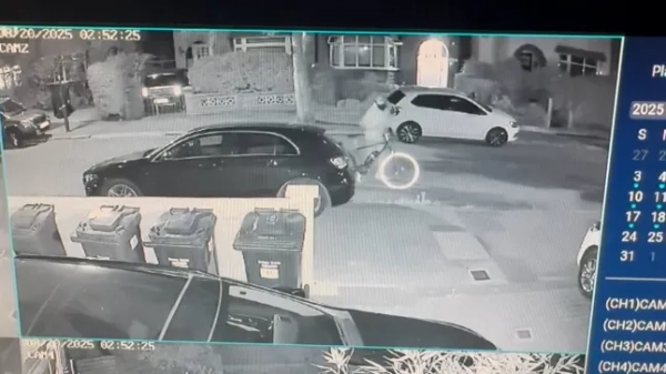 Знімок екрана з камери відеоспостереження, на якому видно чоловіка на велосипеді, який об'їжджає чорний автомобіль, припаркований на житловій дорозі