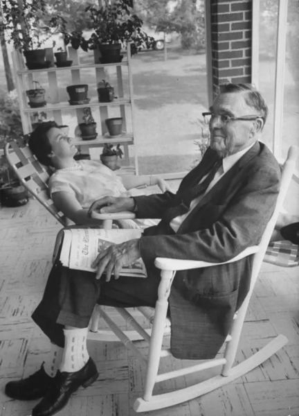 На передньому плані літній чоловік у кріслі-гойдалці з газетою в руках, на задньому плані — молода жінка сидить, відкинувшись у кріслі