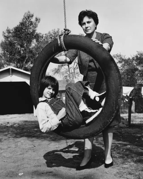 Дівчинка з короткою стрижкою та в комбінезоні сидить на імпровізованих гойдалках із шини, що висить на мотузці. Її хитає жінка з короткою стрижкою, одягнена у сукню