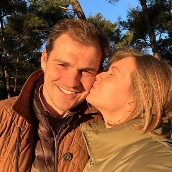 Двоє людей стоять близько одне до одного на передньому плані, одягнені в ватні куртки, одна коричнева, а інша зелена. Вони стоять надворі в лісистій місцевості з високими соснами позаду. Тепле сонячне світло падає на сцену, а блакитне небо видно крізь дерева на задньому плані.