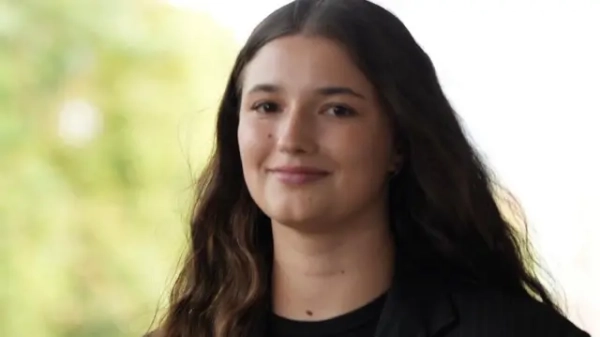 Брюнетка стояла на тлі природи, вона посміхалася в камеру, а її волосся майоріло на вітрі. На ній був чорний піджак у тонку смужку з трояндою збоку.