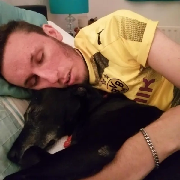 Молодий чоловік спить на ліжку, обіймаючи свого собаку. Він одягнений у жовту футбольну футболку, а лівою рукою обіймає собаку. 