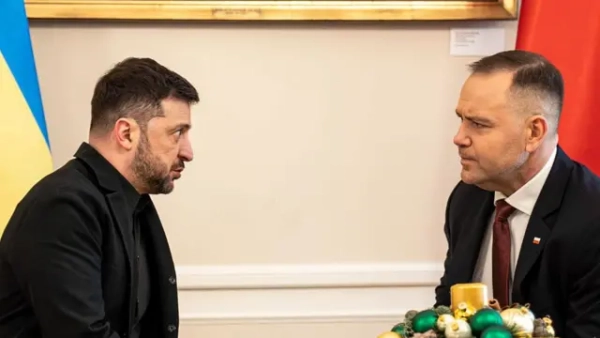 Зеленський і Навроцький сидять навпроти одне одного за столом, позаду видно прапори України та Польщі. Обличчя обох спрямовані одне до одного, брови злегка зведені, губи стиснуті, погляди зосереджені, що створює враження напруженої та уважної розмови.