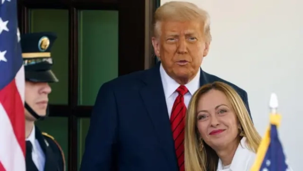Мелоні та Трамп на порозі Білого дому. Трамп – у темному костюмі й червоній краватці – стоїть позаду і щось говорить, дещо примруживши очі. Перед ним дуже близько стоїть усміхнена Мелоні, яка ніби лише на мить обернулася обличчям до камери. Ліворуч частково видно військовослужбовця у формі та кашкеті та прапор США. 