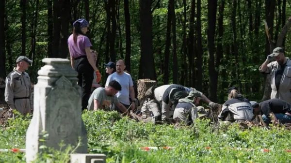 У лісі група людей проводить розкопки на невеликій ділянці землі. Частина людей нахилилася й працює інструментами в ґрунті, поруч стоять інші, спостерігаючи. На передньому плані видно старий кам’яний надгробок, навколо — зелена трава та дерева.