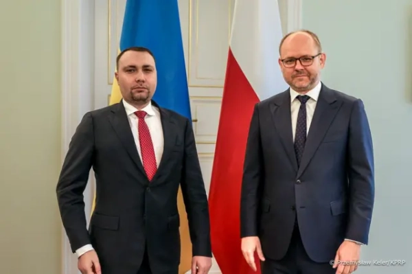Буданов і Пшидач стоять поруч у формальному інтер’єрі на тлі прапорів України та Польщі, одягнені в ділові костюми з краватками. Обоє дивляться прямо в камеру з серйозними, зосередженими обличчями, без помітних емоцій.