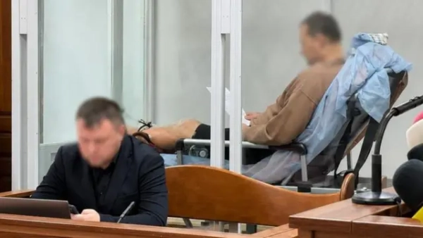 Аварія в Києві: чотирьох загиблих внаслідок наїзду водія, суд його заарештував 8 8 червня, фото із зали суду - на передньому плані сидить чоловік, на задньому - інший чоловік напівлежить на медичній кушетці, їхні обличчя розмиті