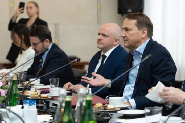 Кілька учасників у діловому одязі сидять перед мікрофонами, документами та напоями. На передньому плані видно пляшки з водою, чашки та папки з документами. Сікорський жестом руки супроводжує виступ або репліку під час обговорення. 