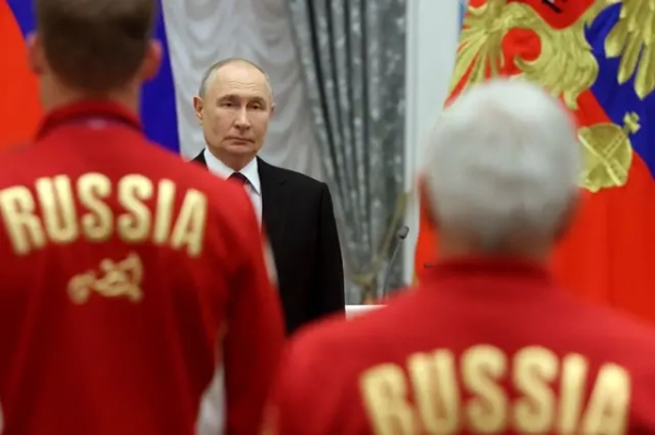 Офіційний захід у президента РФ: на задньому плані стоїть Путін на тлі російського прапора. На передньому плані, спиною до камери, стоять двоє людей у червоних спортивних куртках із золотистим написом «RUSSIA». Композиція кадру акцентує увагу на зв’язку між російською спортивною делегацією та державними символами Росії.