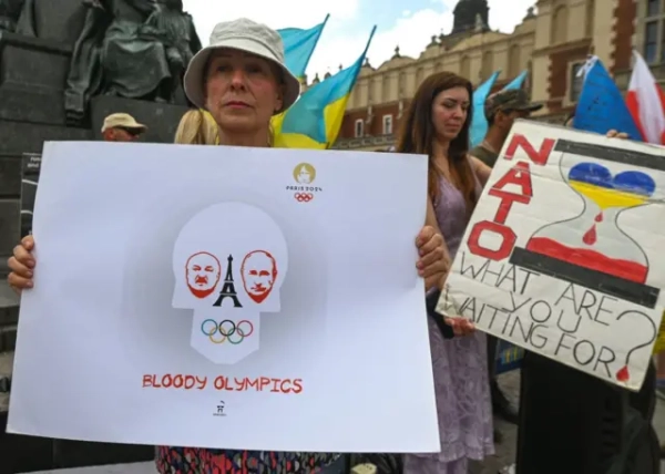 На передньому плані людина тримає великий плакат із написом англійською: «Кривава Олімпіада», олімпійськими кільцями та логотипом Paris 2024. Поруч видно плакат із написом англійською: «НАТО, чого ви чекаєте?» та зображенням пісочного годинника в кольорах українського прапора. На задньому плані майорять українські прапори, видно пам’ятник і будівлі історичного центру міста.