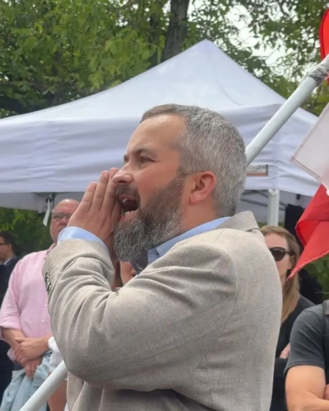 На відкритому заході людина в світлому піджаку стоїть серед натовпу та вигукує щось, приклавши долоню до рота. За спиною видно білий намет і польський прапор. Навколо зібралися учасники заходу, частина з них спостерігає за подією.