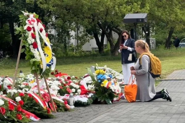 На меморіальному місці вздовж доріжки лежать численні вінки та букети квітів, переважно в біло-червоних кольорах польського прапора. На передньому плані жінка стоїть на колінах біля квітів і вінків, серед яких є композиції в синьо-жовтих кольорах.