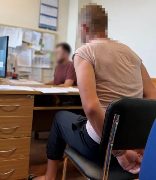 Троє громадян України в Польщі підозрюються у смертельному побитті чоловіка. Посольство України надало коментар щодо справи. 5 У робочому кабінеті на передньому плані людина сидить на стільці біля письмового столу, тримаючи руки, сковані кайданками, за спиною. Обличчя людей на знімку навмисно розмите. На столі лежать документи та папери, ліворуч видно комп’ютерний монітор і тумбу з шухлядами.