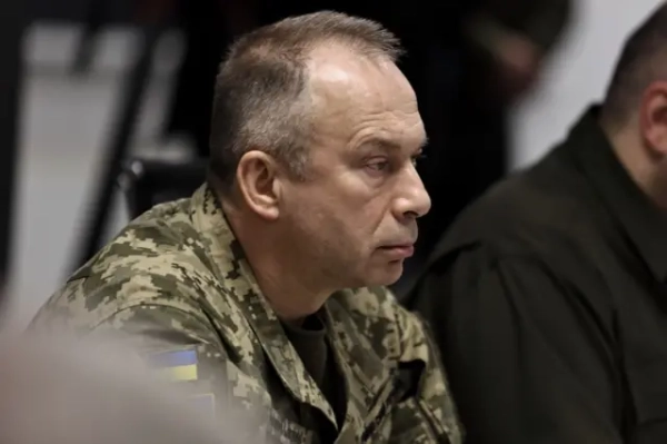 Сирський у піксельній військовій формі сидить за столом під час наради. На рукаві видно нашивку з прапором України. Фото зроблене збоку крупним планом: у фокусі обличчя та верхня частина форми.