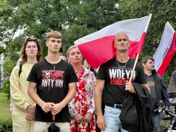 Кілька чоловіків і жінок стоять із польськими прапорами на тлі дерев. На передньому плані – двоє чоловіків у чорних футболках. На одній із них напис: "Анти УПА" та перекреслений портрет Степана Бандери та перекреслений червоно-чорний прапор. Чоловік праворуч тримає великий польський прапор. Усі звернені в один бік, слухають виступ.