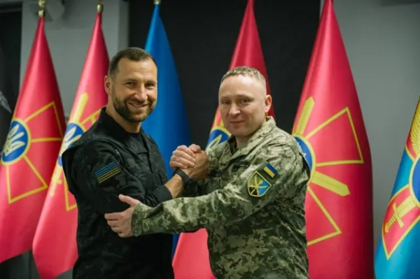 Зеленський ухвалив рішення: Драпатого призначено головнокомандувачем, Скибюк – очільником Генштабу. 5 Двоє чоловіків у військовій формі стоять у приміщенні та тиснуть одне одному руки, охопивши їх обома долонями. Один одягнений у темну форму, інший — у піксельний камуфляж із нашивкою прапора України на рукаві. На задньому плані розташовано кілька червоних прапорів із військовою символікою та один синій прапор.