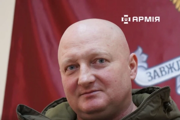 Зеленський ухвалив рішення: Драпатого призначено головнокомандувачем, Скибюк – очільником Генштабу. 6 Портрет крупним планом чоловіка середнього віку на тлі червоного прапора. Людина одягнена в темно-оливкову куртку. На задньому плані частково видно емблему та фрагмент напису на стрічці. Фото добре освітлене, основна увага зосереджена на обличчі в центрі кадру.
