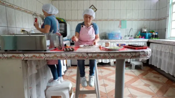 Марія Луїса працює біля кухонної стільниці, сидячи на високому стільці. На ній рожевий фартух та сітка для волосся. Позаду неї сестра працює, стоячи на сходинці, щоб дістати до стільниці.