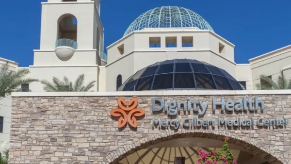 Будівля медичного центру Dignity Health Mercy Gilbert Medical Center. На передньому плані видно кам’яну стіну з назвою лікарні, позаду — світла будівля з великими скляними куполами й пальмами навколо.