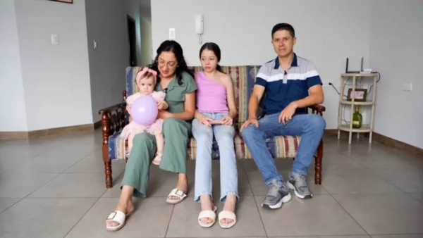 Каміла сфотографована разом із родиною, яка сидить на дивані у вітальні з кахельною підлогою. Вона в рожевій сукні та обручі для волосся, тримає в руках фіолетову повітряну кульку. Вона сидить на колінах у матері з довгим темним волоссям та окулярами, одягненої в зелений комбінезон. Її сестра сидить посередині в фіолетовому топі з відкритими плечима та джинсах поруч із батьком у синій футболці-поло та джинсах.
