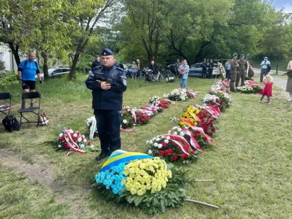 На галявині розкладені десятки вінків і квіткових композицій, переважно в польських національних кольорах. На передньому плані лежить вінок із жовтих і блакитних жоржин. Біля нього стоїть працівник поліції, а позаду видно поодиноких учасників церемонії.