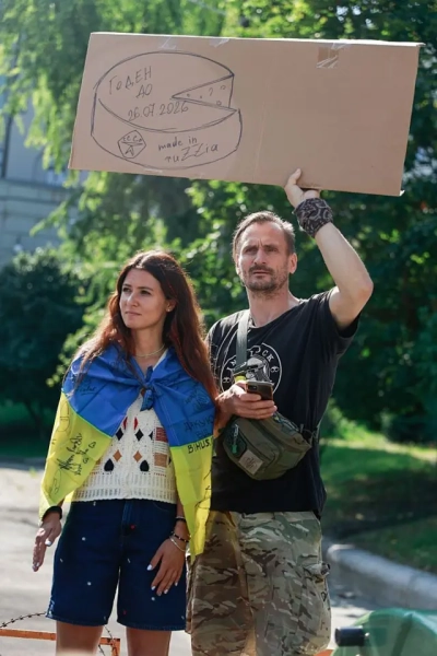 Чи чинять командири тиск на військових через протести з картону? Офіс президента та "Мадяр" дали відповідь 6 Жінка та чоловік на протесті. Чоловік у мілітарі-штанях тримає над головою великий картонний плакат із намальованим кругом сиру та написами, зокрема "made in ruzzia" і "годен до 26.07.2026". Жінка загорнута в український прапор із численними підписами.