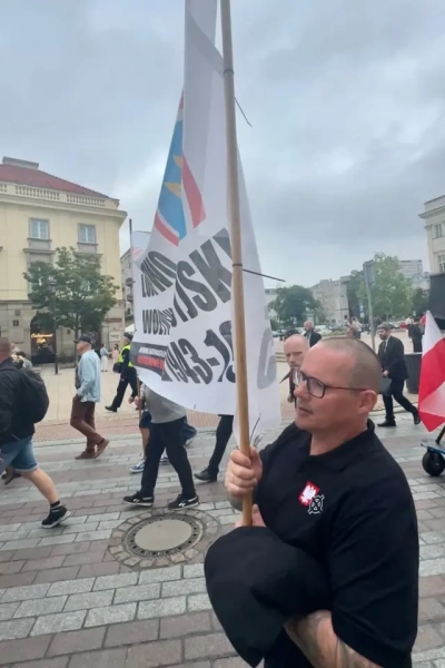 Чоловік у темній футболці та окулярах тримає високий банер із перекресленим тризубом на дерев’яному держаку. На грудях видно два невеликі символи: червоно-білий значок із польським орлом та білий кельтський хрест (вертикальний хрест, обведений колом). Навколо йдуть учасники зібрання, поруч видно польський прапор, історичні будівлі та перехожих.