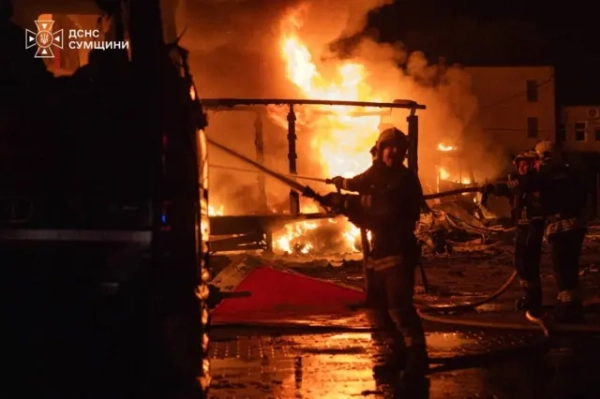 Рятувальники ДСНС ліквідовують масштабну пожежу в нічний час. На задньому плані видно потужне полум’я, яке охопило будівлю або промисловий об’єкт, та густий дим. Пожежники працюють зі шлангами біля пожежного автомобіля, а мокре покриття відбиває яскраве світло вогню.