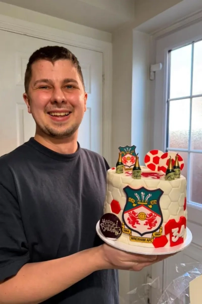 Джош у темній футболці тримає святковий торт із символікою Wrexham FC. Він усміхається й дивиться в камеру.