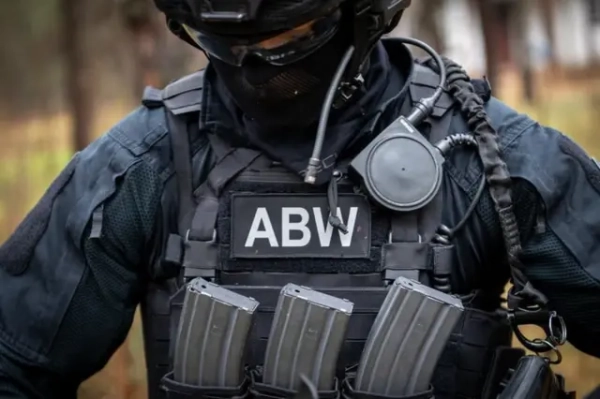 Співробітник польської спецслужби в чорному тактичному спорядженні, шоломі та захисній масці. На бронежилеті розміщена нашивка «ABW» (Агентство внутрішньої безпеки Польщі), а також магазини до зброї та засоби звʼязку.