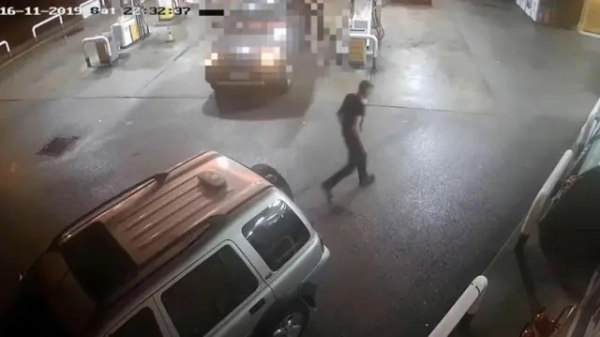 На записі з камери спостереження видно, як людина йде мокрим майданчиком біля автозаправки вночі. Поруч припарковані кілька автомобілів біля паливних колонок, у верхньому лівому куті видно позначку часу.