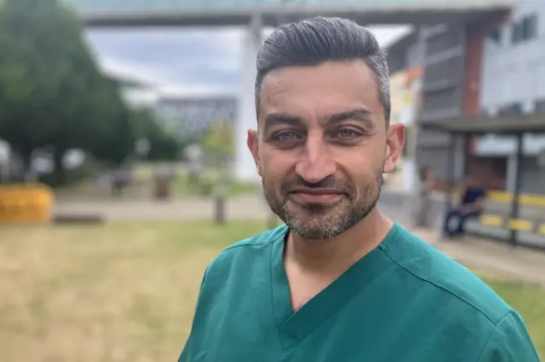 The image shows a close-up portrait of a person standing outdoors, wearing teal V-neck medical scrubs. The photograph is sharply focused on the person in the foreground, while the background is blurred with a shallow depth-of-field effect. In the background, there appears to be a grassy area, trees, a modern building, and a bus stop or shelter with a few people nearby. 