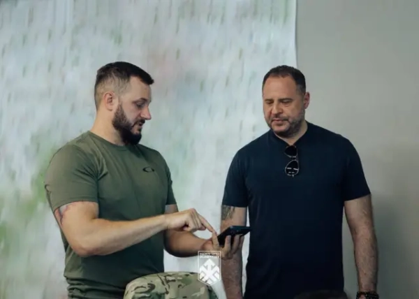 Єрмак та Хартія. Як інтеграція корпусу до програми "Адвокат+" викликала хвилю дискусій 6 Двоє чоловіків стоять у приміщенні та дивляться на екран смартфона, який один із них тримає в руці. На передньому плані частково видно військове спорядження, а на зображенні є логотип підрозділу "Хартія".