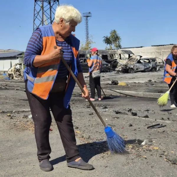 На передньому плані літня жінка в помаранчевому сигнальному жилеті змітає уламки з асфальту мітлою. Навколо розкидані обгорілі фрагменти та сміття. На задньому плані видно кілька сильно пошкоджених і вигорілих автомобілів, а також інших людей, які долучилися до прибирання.