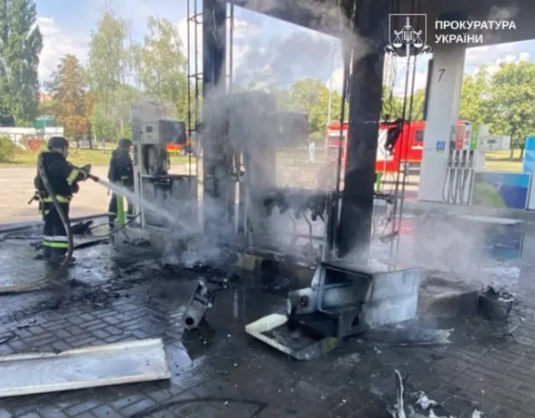 Пошкоджена АЗС після пожежі. Два пожежники гасять згорілу колонку, з якої ще підіймається густий дим. Навколо лежать обгорілі уламки конструкцій, а на задньому плані видно червоний автомобіль ДСНС.