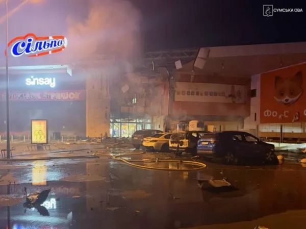 У нічний час біля торговельного центру видно значні пошкодження будівлі: частина фасаду та даху зруйнована, зсередини піднімається дим. Перед входом стоять кілька автомобілів, навколо розкидані уламки, а на мокрому асфальті лежать пожежні рукави. На фасаді помітні вивіски магазинів «Сільпо», Sinsay і дитячого магазину. 
