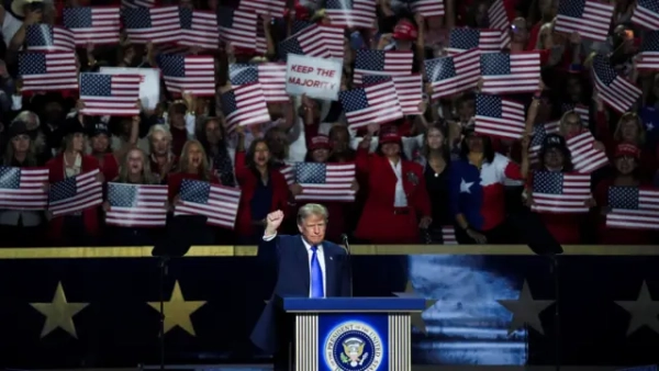 Трамп обіцяє $5 тис кожному американцю за успіх республіканців 4 На великій сцені під час політичного заходу Трамп виступає за трибуною, піднявши стиснутий кулак. Позаду нього зібрався натовп, який тримає десятки прапорів США та плакати.